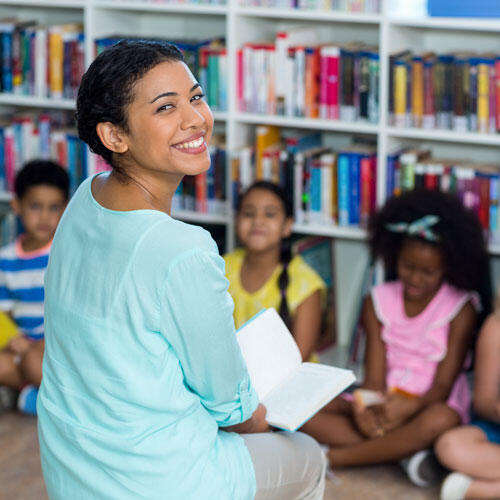 An elementary school teacher sitting on the floor with her students looking back at the camera